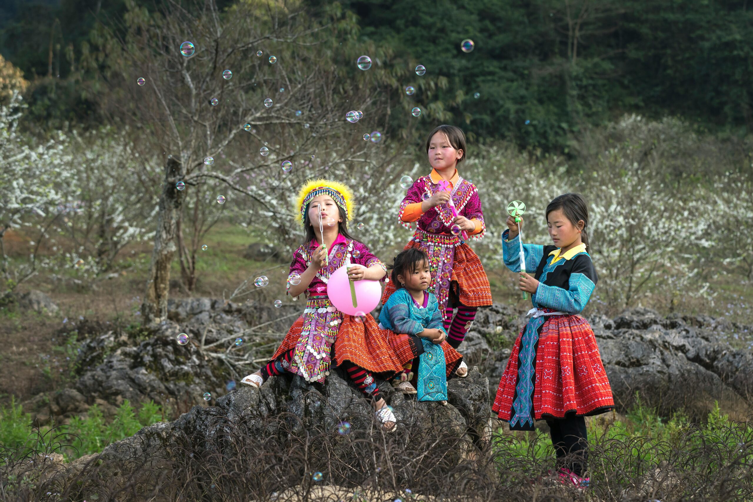 pexels-photo-2171423-2171423 Four children in colorful traditional attire play with soap bubbles outdoors, surrounded by a scenic landscape.